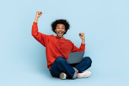 Afroamerikaner, der mit Laptop auf dem Boden sitzt, schlägt triumphierend die Luft an