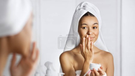 Mignonne afro-américaine fille appliquant de la crème sur son visage dans la salle de bain, regardant dans le miroir, réfléchissant
