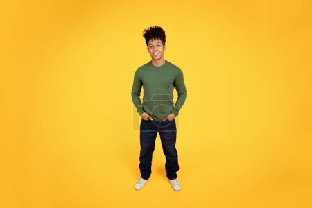 Foto de Un joven negro feliz vestido con ropa casual se para con confianza con las manos en los bolsillos sobre un llamativo fondo amarillo - Imagen libre de derechos