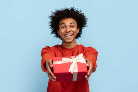 Téléchargez les photos : Homme afro-américain joyeux tend une boîte-cadeau rouge avec un ruban blanc, l'air amical - en image libre de droit
