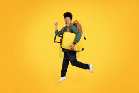 Young energetic african american guy with a big smile running and holding a yellow suitcase against a yellow background
