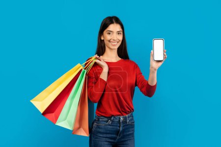Photo for Woman holding multiple shopping bags and showing blank cell phone, happy female recommending new app for purchases, standing on blue studio background, mockup - Royalty Free Image
