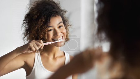 Foto de Mujer afro joven sonriente con cepillo de dientes limpiando los dientes en el baño. Concepto de higiene oral matutina - Imagen libre de derechos