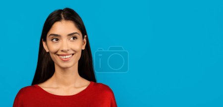 Téléchargez les photos : Portrait d'une jeune femme souriante portant un pull rouge, les cheveux baissés, debout sur un fond de studio bleu vif, regardant de côté l'espace de copie, panorama - en image libre de droit