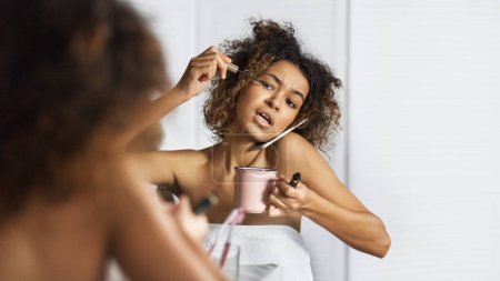 Afro-american girl in hurry put on makeup, drinking coffee and talking by phone simultaneously in front of mirror in bathroom. Crazy morning concept