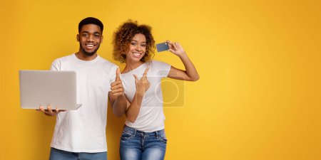 Photo for A young smiling African American couple stands against a yellow backdrop, man holds a laptop while woman gestures thumbs up. - Royalty Free Image