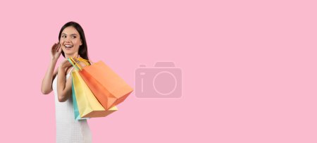 Téléchargez les photos : Une dame européenne joyeuse porte plusieurs sacs à provisions, suggérant une vente d'été, faisant appel à la génération Z sur un fond rose isolé, espace de copie - en image libre de droit