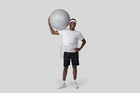 Téléchargez les photos : Contenu L'homme afro-américain en tenue d'entraînement tient une balle de gym à ses côtés et sourit, sur fond de studio ordinaire - en image libre de droit