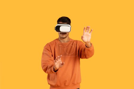Téléchargez les photos : Un gars est immergé dans la réalité virtuelle, portant un casque VR et gesticulant avec leurs mains sur un fond orange vif, visage obscurci pour la vie privée - en image libre de droit