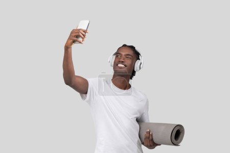 Junger afroamerikanischer Mann mit Kopfhörer auf dem Handy, um ein Selfie zu machen, hält Yogamatte auf weißem Hintergrund