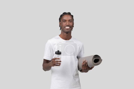 Téléchargez les photos : Souriant athlète afro-américain tient une bouteille d'eau et tapis de yoga, prêt pour l'exercice sur fond blanc - en image libre de droit