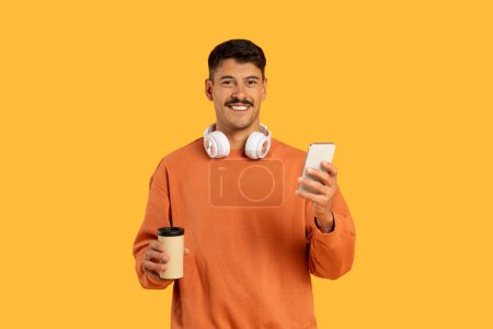 Photo for Man with moustache in an orange sweater with headphones, a coffee cup, and a smartphone, on an orange backdrop - Royalty Free Image
