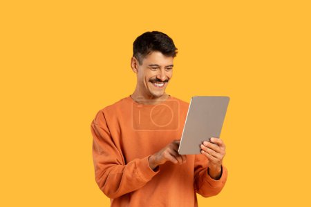 Glücklicher Mann im orangefarbenen Pullover, der auf einen Tablet-Bildschirm mit freudigem Gesichtsausdruck auf gelbem Hintergrund blickt