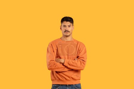 A stern-looking man with moustache stands with arms crossed on a solid yellow background, copy space