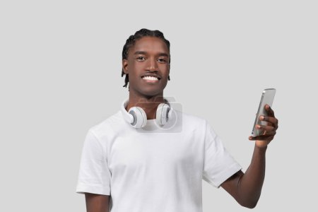 Feliz joven afroamericano sonriendo mientras usa su teléfono inteligente, usando auriculares sobre fondo blanco