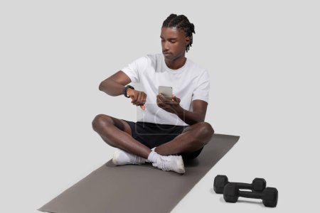 Focused African American guy athlete sits on a yoga mat looking at his smartwatch, with dumbbells nearby in a neutral studio