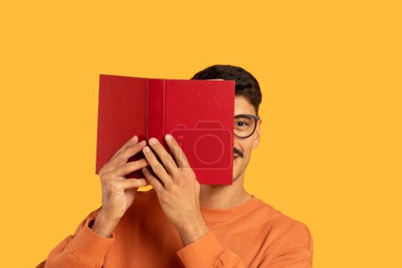 A man hides his face behind a red book against a vibrant orange backdrop, invoking curiosity and mystery