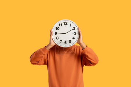 Téléchargez les photos : Un gars en pull orange recouvre son visage d'une horloge murale, symbolisant les concepts de temps et de délais - en image libre de droit