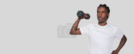 Foto de Fit chico afroamericano posa casualmente con una mancuerna sobre el hombro, transmitiendo confianza y fuerza en un entorno minimalista - Imagen libre de derechos