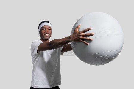 An athletic young African American man in active clothing demonstrates a chest pass with a large exercise ball against a plain backdrop