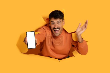 Foto de Hombre emocionado rasga a través de fondo de papel naranja, presentando la pantalla del teléfono inteligente con un gesto animado - Imagen libre de derechos