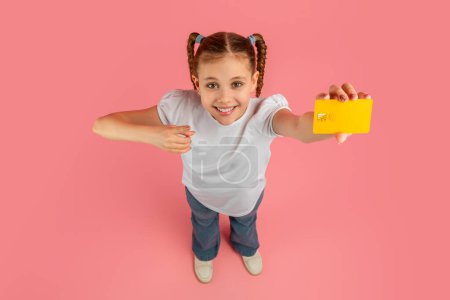 Photo for A cheerful young girl with braided hair smiling and showing yellow credit card on a pink background - Royalty Free Image
