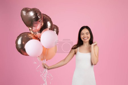 Foto de Una dama sonríe mientras tira de los hilos de globos en forma de corazón, aislados sobre fondo rosa del estudio - Imagen libre de derechos