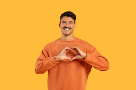Téléchargez les photos : Homme joyeux formant un symbole de coeur avec ses mains, exprimant amour ou affection sur une toile de fond jaune - en image libre de droit