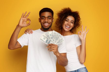 Foto de Gran inversión. Feliz africano hombre y mujer gesto OK signo con un montón de dinero, fondo de estudio naranja - Imagen libre de derechos
