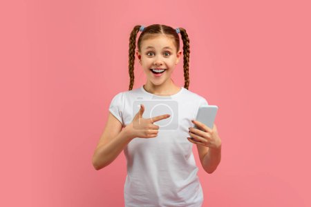 Chica joven con el pelo trenzado señala con entusiasmo a su teléfono inteligente sobre un fondo rosa, expresando sorpresa y deleite