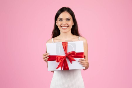 Foto de A lady showcases a gift wrapped with a red ribbon, a universal symbol of giving during summer, isolated on pink - Imagen libre de derechos