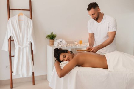 A masseur employs a wooden massage tool on a clients African American woman back during a spa procedure