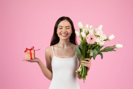 Téléchargez les photos : Une dame souriante tient un beau bouquet d'été et un cadeau enveloppé, incarnant l'enthousiasme de la génération Z. Isolé sur un fond rose - en image libre de droit