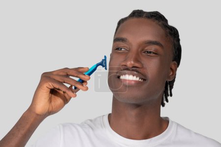 Close-up of a cheerful African American man using a blue razor to shave his face, depicting guy grooming