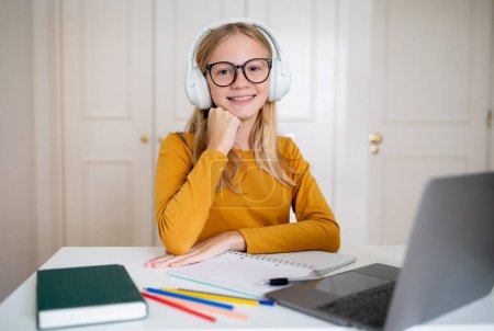 Téléchargez les photos : Une adolescente joyeuse portant des lunettes et des écouteurs assis à un bureau avec un ordinateur portable et du matériel d'étude dans une pièce lumineuse à la maison, adolescente souriante à la caméra - en image libre de droit