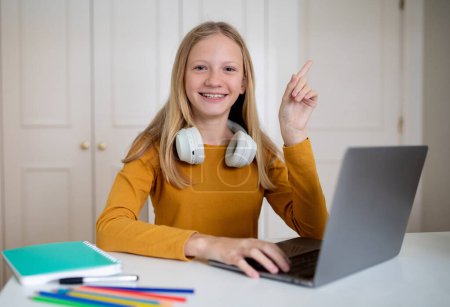 Téléchargez les photos : Une adolescente de contenu avec un casque autour du cou lève la main en utilisant un ordinateur portable, étudier à la maison, ce qui indique une interaction e-learning. - en image libre de droit