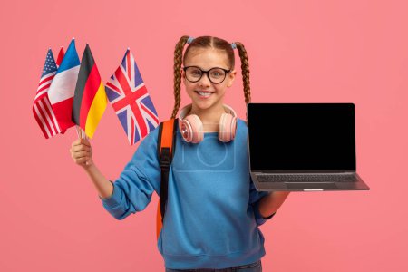 Mädchen mit Brille und Zöpfen hält einen Laptop mit leerem Bildschirm und mehreren internationalen Flaggen, die die globale Verbundenheit symbolisieren