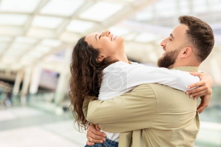 Foto de Un hombre y una mujer abrazándose felizmente en una terminal del aeropuerto iluminada por el sol después de la llegada, pareja encantadora feliz de verse, compartiendo momento romántico, primer plano - Imagen libre de derechos