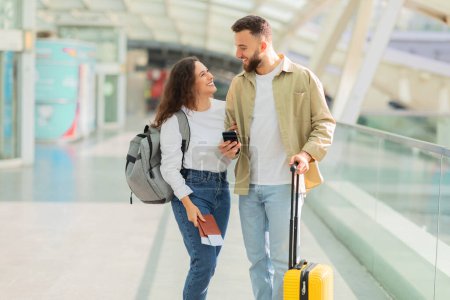 Photo for A man and woman are walking through an airport with their luggage and smartphone in hand, happy spouses looking at each other, enjoying travelling together - Royalty Free Image