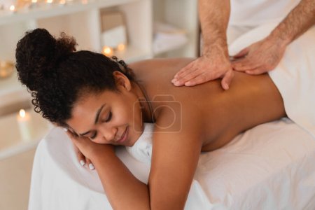 Téléchargez les photos : Afro-Américaine jeune femme couchée face contre terre profitant d'un massage du dos professionnel dans un cadre spa serein - en image libre de droit