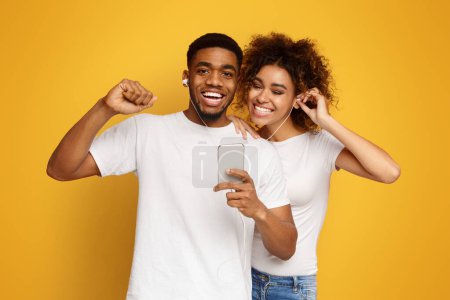 Photo for Cheerful black couple sharing headphones, enjoying favorite music on smartphone, orange background - Royalty Free Image