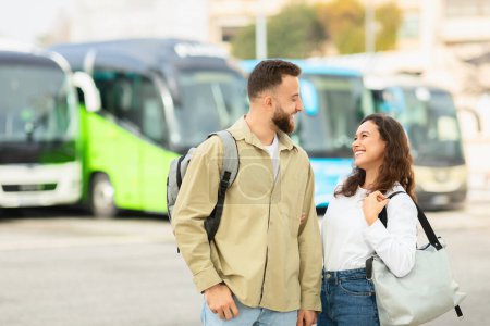 Photo for Happy man and woman with backpacks smiling at each other, ready to board a bus for a trip, enjoying travelling together - Royalty Free Image