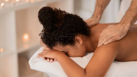Afroamerikanerin in ruhiger Wellness-Umgebung, die eine entspannende Schultermassage von einem männlichen Masseur erhält