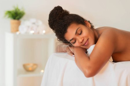 Téléchargez les photos : Femme afro-américaine aux yeux fermés embrasse un oreiller, apparaissant serein et détendu sur une table de massage spas - en image libre de droit