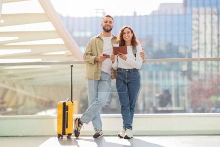 Un hombre y una mujer sonrientes sosteniendo pasaportes pasean casualmente por un aeropuerto, listos para viajar con una maleta amarilla.