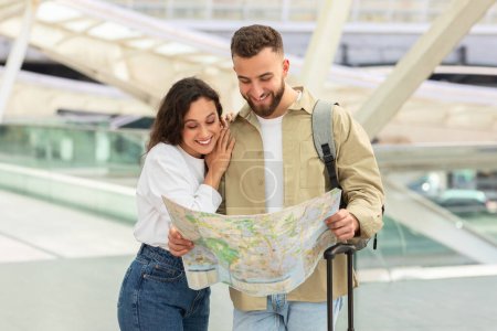 Foto de Sonriente joven hombre y mujer mirando el mapa juntos, de pie en el moderno centro de transporte o aeropuerto, feliz pareja comprobando la ruta de viaje, espacio libre - Imagen libre de derechos