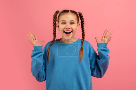 Téléchargez les photos : Une jeune fille enthousiaste lève les mains dans l'excitation portant un pull bleu sur un fond rose - en image libre de droit