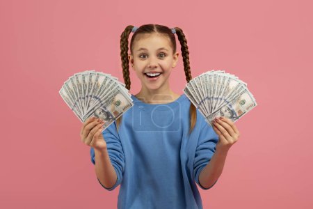 A delighted girl in a blue sweater holding a fan of dollar bills with a thrilled expression on a pink background