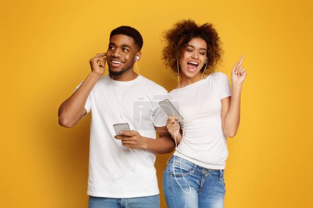 Téléchargez les photos : Couple émotionnel afro-américain à l'écoute de musique dans les écouteurs, fond studio orange - en image libre de droit