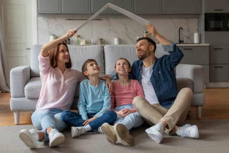 Photo for Smiling family sitting on floor makes a house shape with carton card, symbolizing unity and home, secure environment for kids - Royalty Free Image